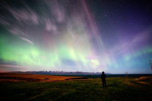 A man watches the Aurora Borealis by a derelict farm building, Stars shine brightly within the surrounding darkened sky. North-Eastern Iceland